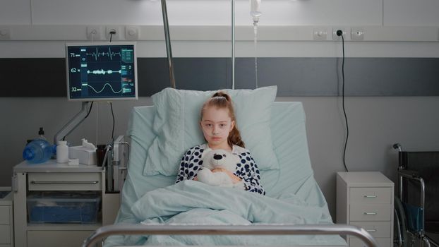 Portrait of hospitalized sick girl child patient holding teddy bear resting in bed during medical consultation in hospital ward. Alone kid wearing oxygen nasal tube waiting for disease examination