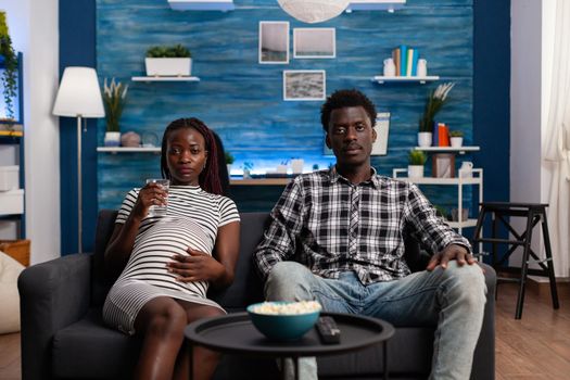 POV of black couple expecting baby watching TV together on living room couch. African american people looking at camera and television while pregnant woman holding glass of water