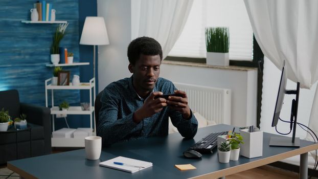 Cheerful happy black man playing video games on his phone in home office while taking a break from remote working from home. African american male having fun, enjoying enternainment on mobile phone