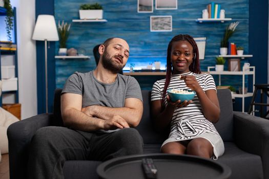 POV of interracial couple enjoying activity together at home. Mixed race partners smiling and watching television in living room, looking at camera and eating popcorn snack at TV