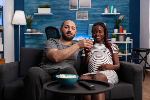 POV of interracial couple with pregnancy watching movie on television in living room. Multi ethnic people expecting baby and looking at camera while having snacks and water at home