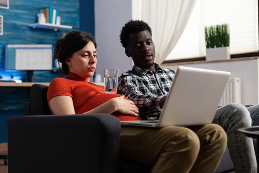 Pregnant interracial couple relaxing and using technology. Multi ethnic partners expecting baby, caucasian woman holding modern laptop and black father of child looking at device screen