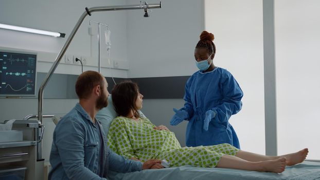 Wife expecting child sitting in hospital ward with husband waiting for delivery preparations at maternity clinic. Young caucasian couple talking to african american nurse about childbirth