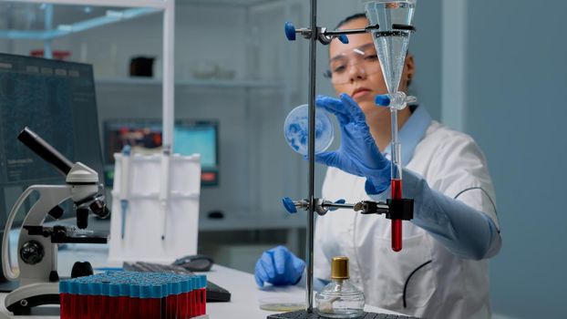 Microbiology woman studying petri dish in laboratory at clinic, having chemical equipment. Doctor doing medical research with vacutainers, test tube, micropipette and microscope on desk