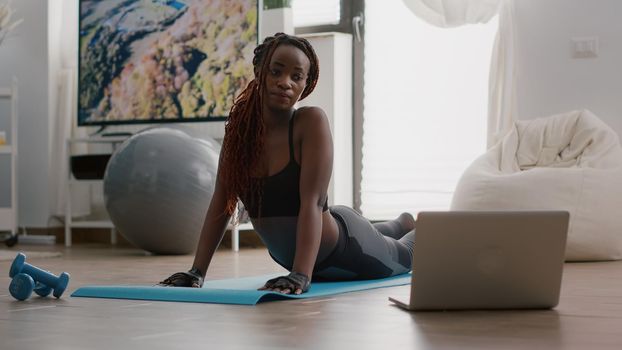 Flexible black woman practicing sport during yoga morning workout sitting on fitness map in living room. Adult stretching body muscles watching training aerobic video using laptop computer