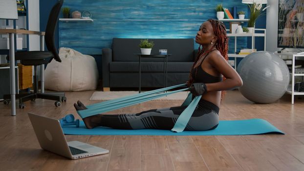 Athetic black woman in sportswear watching aerobic online sport workout on laptop working body muscle using fitness band enjoying healthy lifestyle. Slim adult sitting on yoga map in living room