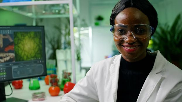 Close up of african biologist doctor wearing medical equipment smiling at camera while sitting in microbiology laboratory. Specialists researching genetic mutation developing dna test