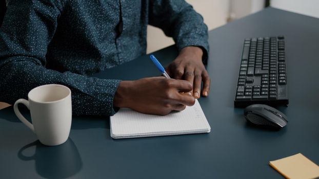 Close up of african american black person taking notes on notepad using a pen. Male adult hands of remote worker writing text on white paper