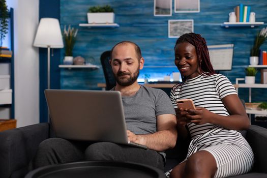 Modern interracial couple using devices and technology at home. African american woman holding smartphone while caucasian man working on digital laptop. Mixed race people with gadgets