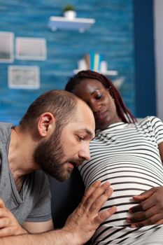 Close up of caucasian father of baby looking at belly and holding hand while pregnant black woman smiling. Interracial couple with pregnancy expecting child and relaxing at home