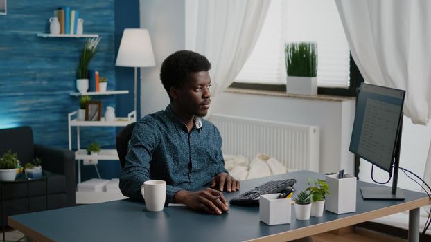 Black african american freelancer taking notes on notepad and looking into computer, remote working from home computer user