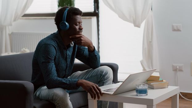 Authentic smiling african american man using laptop with headphones on, working from home or freelancer listening webinar or online courses. Taking internet webinar from home office