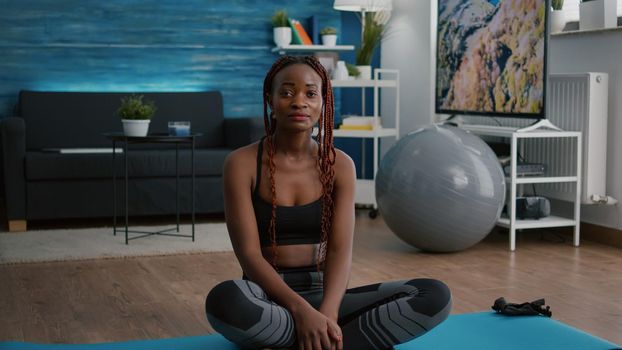 Portrait of athlete fit woman looking into camera while sitting in lotus position on yoga map during morning fitness workout in living room. Flexible adult enjoying healthy lifestyles