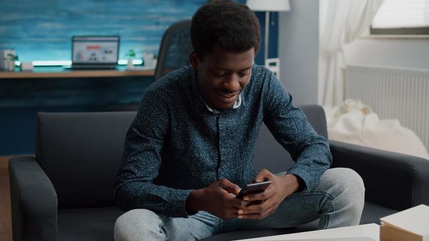 Black guy in his living room using phone to browse social media, internet connection and communication device. African american man working from home, remote worker in wirelles home