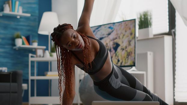 Athlete black woman training on yoga map in living room standing in side plank watching fitness video using laptop computer. Adult in sportwear streching muscle during strech gymnastics