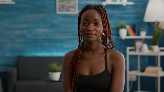 Portrait of athlete with black skin looking into camera while siting on aerobics ball during morning workout in living room. Strong adult practicing gymnastics warming before aerobics