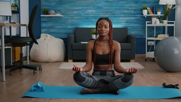 Portrait of black woman sitting in lotus position on floor doing morning breathing workout in living room. Athlet slim fit adult wearing sportwear stretching muscle during body gymnastics
