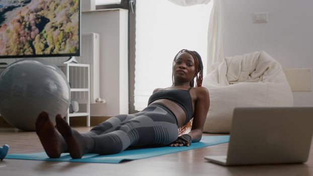 African slim woman morning workout in living room doing scissors exercise on yoga map watching aerobic online class using laptop. Athlete person in sportswear stretching leg muscle