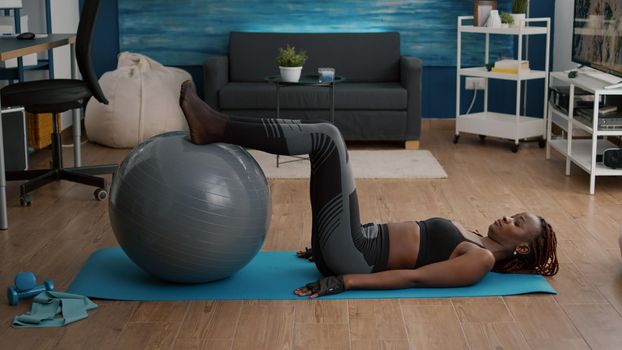 Athletic woman with black skin working abs on swiss ball in living room during morning workout in living room. Strong sllim fit adult in sportsweartraining body muscle exercising pilates pose