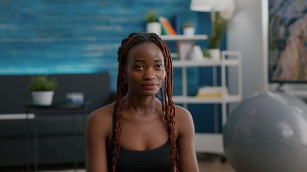 Black woman trainer sitting in lotus position on yoga mat on floor with closed eyes meditating during morning fitness bodycare workout. Adult doing breathing exercises enjoying healthy lifestyle