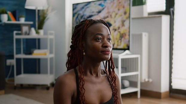 Flexible slim woman with black skin practicing respiratory exercise during morning fitness workout in living room sitting on yoga map. Athlete adult stretching body muscles enjoying healty lifestyles