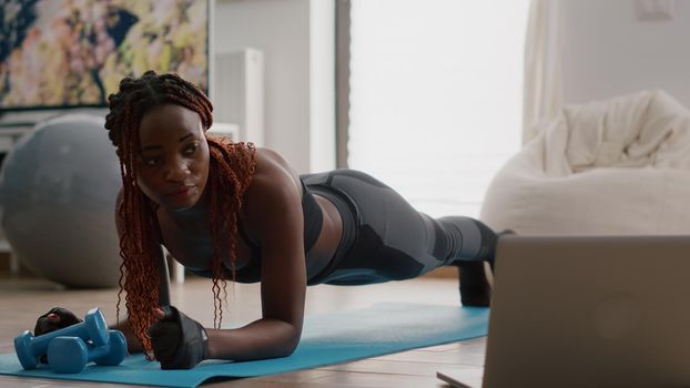 Afro american woman with sportswear stretching yoga exercises on fitness map in living room watching online pilates video using laptop computer. Healthy person doing morning gymnastics