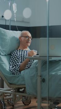 Elder sick patient using laptop in hospital ward bed to talk to relatives about health problems. Old man on videocall waiting for healing treatment to cure pain from surgery and disease