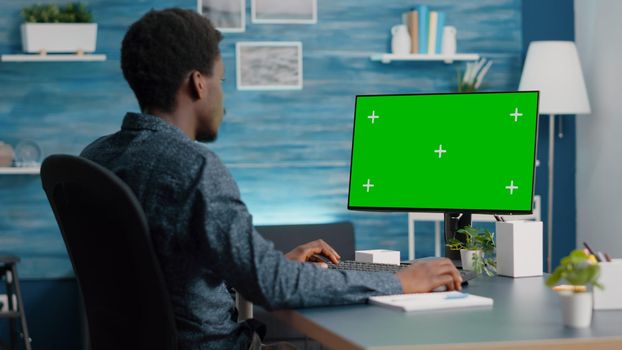African american man using and typing on mockup computer with green screen. Computer user on isolated chroma mock up display in living room, bright house