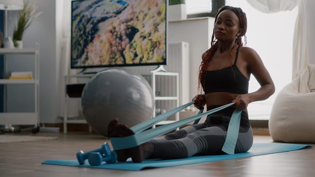 Athlete with black skin in sportswear exercising body muscle using fitness elastic enjoying healthy lifestyle sitting on yoga map in living room. Fit woman working at wellness warming before workout