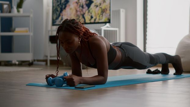 Athlete african in sportwear doing fitness exercices putting cronometer on phone sitting on yoga map in living room. Flexible woman stretching muscle body enjoying healthy lifestyle