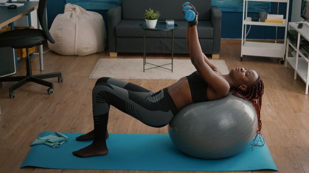 Slim athletic woman with dark skin working body muscle doing morning fitness exercices with dumbbells. Fit adult sitting on fitness swiss ball during yoga sport workout in living room