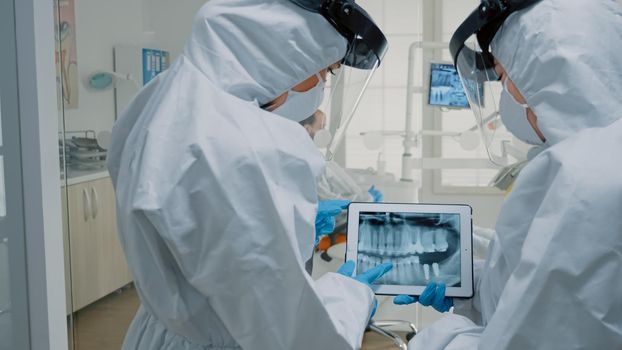 Professional dentists using technology for oral examination while wearing protection suits at dental clinic. Stomatology doctors discussing and looking at x ray scan on modern tablet