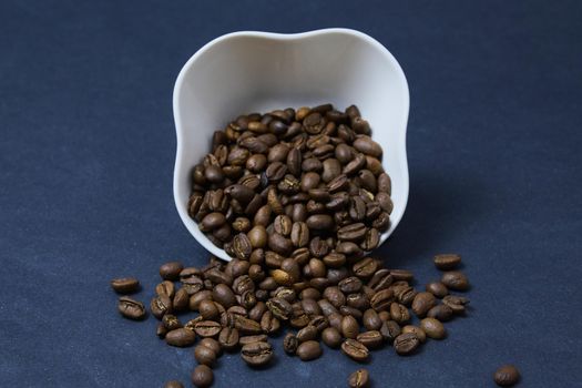 brown coffee beans roasted on a black background spilled out of an upturned white cup on the table