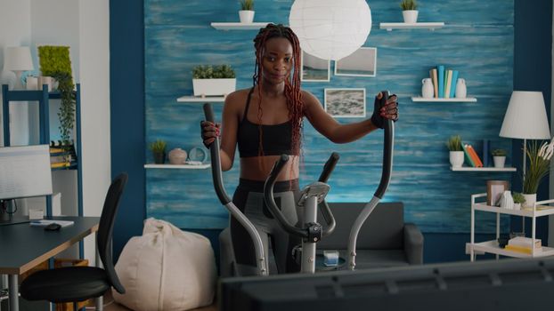 Black woman athletic training on elliptical bike practicing cardio sport watching aerobic video on tv during bodyweight morning routine in living room. Trainer enjoying healthy lifestyle