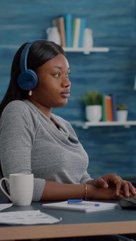 Student with black skin having headphone puts listening online univeristy course using elearning platform sitting at desk in living room. African american woman working remote from home