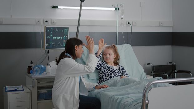 Pediatrician woman doctor relaxing while playing high five with sick girl patient during healthcare examination in hospital ward. Happy little child relaxing in bed interacting with pediatric