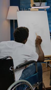 Disabled african american man working on drawing of vase with pencil and white canvas on easel in workshop room. Black artist in wheelchair creating authentic fine art masterpiece