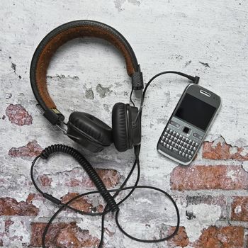 the overhead headphones are connected by a cable to an old push button phone lying on a brick wall with fallen plaster flat lay