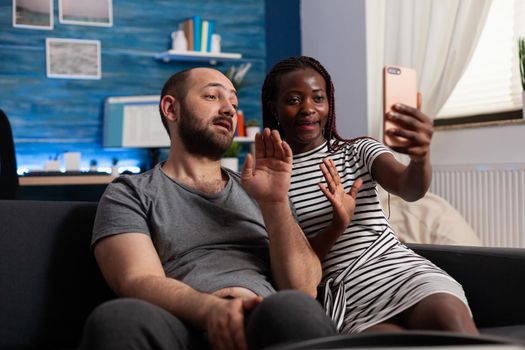 Interracial couple waving at camera while using video call communication. Multi ethnic people holding modern smartphone with online remote conference on internet connection at home.