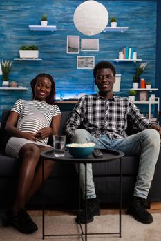 African american couple with pregnancy watching television at home. Young people expecting child while looking at camera and having popcorn. Black pregnant woman and father of baby