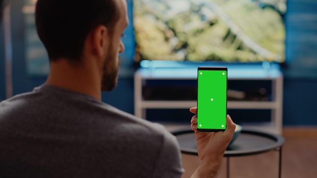 Person with modern smartphone looking at green screen vertical background in living room. Caucasian man with chroma key and isolated media display for mockup copy space template