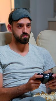 Close up of man with eye sleep mask playing videogames with joystick for gaming competition. Caucasian male sitting in front on television on sofa late at night in kitchen