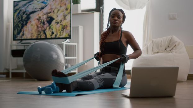 Fit black woman in sportswear looking at online workout on laptop working body muscle using fitness elastic enjoying healthy lifestyle. Athlete adult sitting on yoga map in living room