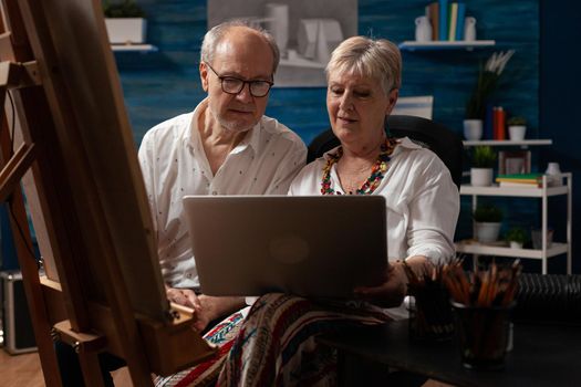 Caucasian elder couple looking at laptop with technology for fine art masterpiece in workspace. Senior people with artistic hobby creating drawing with inspiration from modern gadget