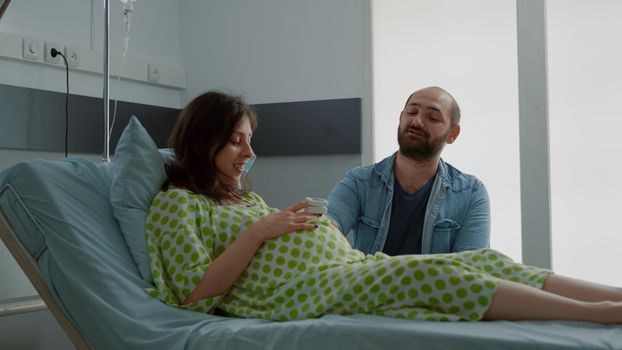 Young family talking about childbirth in hospital ward at maternity clinic. Caucasian pregnant woman laying in bed being happy about delivery. Husband sitting with wife for birth assistance