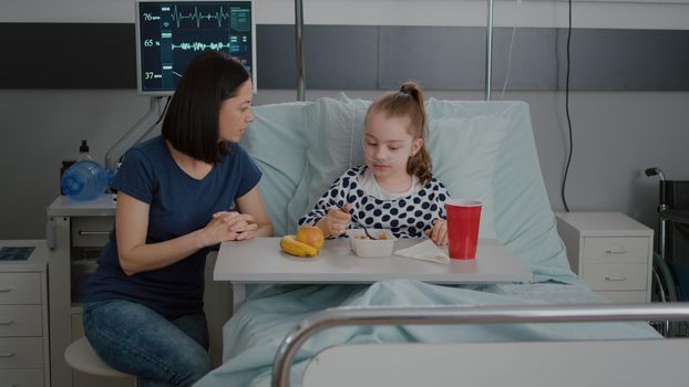 Mother sitting with sick child patient while eating healthy food lunch in hospital ward during sickness examination. Hospitalized girl resting in bed waiting for medication treatment after surgery