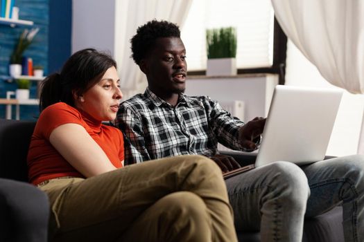 Interracial couple looking at modern laptop computer in living room. African american man holding digital gadget while caucasian woman watching device screen. Mixed race people
