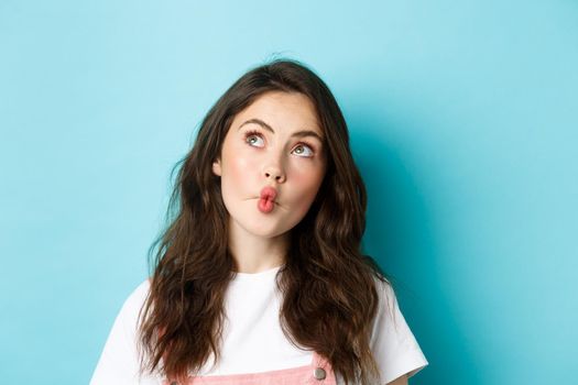 Close up portrait of funny young woman with clean skin and curly hairstyle, pucker lips thoughtful, looking at upper left corner dreamy, imaging something, standing over blue background.