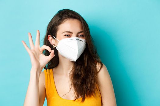 Covid-19, coronavirus and social distancing. Young positive woman in respirator showing okay sign, standing against blue background.