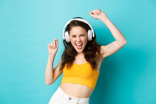 Young woman in summer clothes listening music, wearing headphones and singing along favorite song, dancing in earphones, standing against blue background.